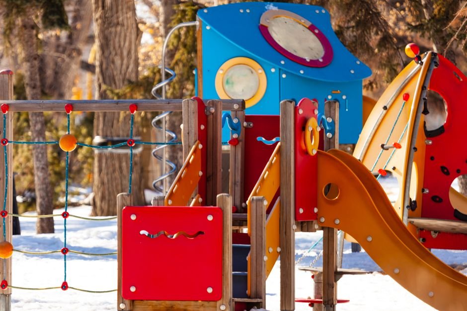 Šviesūs žaidimų aikštelės įrenginiai apsnigtame parke, iliustruojantys priežiūros svarbą žiemą