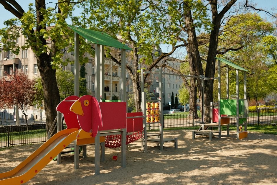 Spalvinga vaikų žaidimų aikštelė su supynėmis ir kalneliais, apsupta medžių ir pastatų
